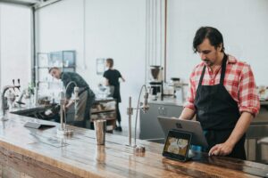 A café manager in a checked shirt and apron uses a tablet at a wooden counter while colleagues work in the background.